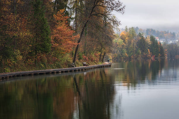 Fiery foliage leans over the tranquil waters of Lake Bled, where a quiet path invites slow steps. Mist lingers in the distance, adding a touch of mystery to the serene scene.