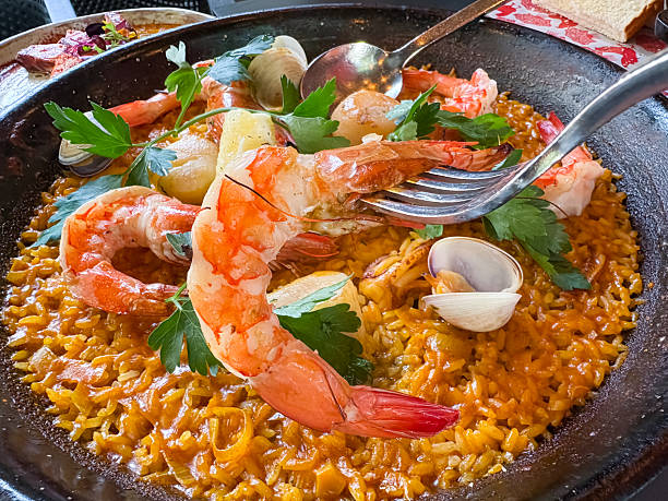 Photo of Spanish seafood paella in typical paellera, with slices of lemons, mussels and clams shells, fork and spoon, and a place for text, shot from above on a teal texture