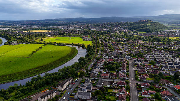 The image captures a sweeping aerial view of Stirling, Scotland, where the River Forth winds gracefully through lush green fields and suburban residential areas. In the distance, the historic Stirling Castle dominates the hilltop, highlighting the city’s medieval heritage and strategic importance in Scottish history. The blend of natural landscapes, modern housing, and historic landmarks reflects the unique character of Stirling as a place where past and present coexist. Known as the “Gateway to the Highlands,” Stirling holds cultural and historical significance, making it a key destination for heritage tourism in Scotland.
