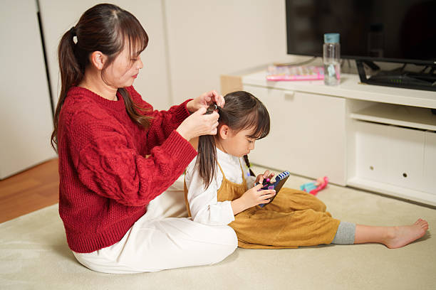 Mother braiding her young daughter’s hair at home while the child plays on a smartphone. Cozy family moment showing daily routine, bonding time, childcare, parenting, and calm indoor lifestyle. Warm domestic atmosphere capturing modern family life and simple everyday interactions.