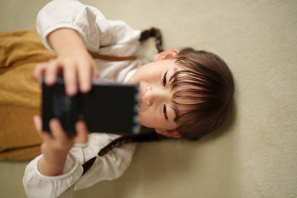 Close-up of a smiling young girl holding a game console while lying on the floor. Child enjoying game time in a relaxed home environment, showing modern childhood, digital entertainment, technology use, and joyful everyday moments.