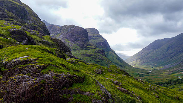 This image presents an aerial perspective of the famous Three Sisters of Glencoe in the Scottish Highlands. The dramatic mountain ridges rise steeply above green valleys, with cascading waterfalls and streams flowing through the rugged landscape. Clouds and mist enhance the moody atmosphere, emphasizing the raw beauty and wilderness of the Highlands. Known as one of Scotland’s most iconic landmarks, the Three Sisters attract hikers, photographers, and travelers seeking dramatic scenery, cultural heritage, and outdoor adventure in a remote and untamed environment.