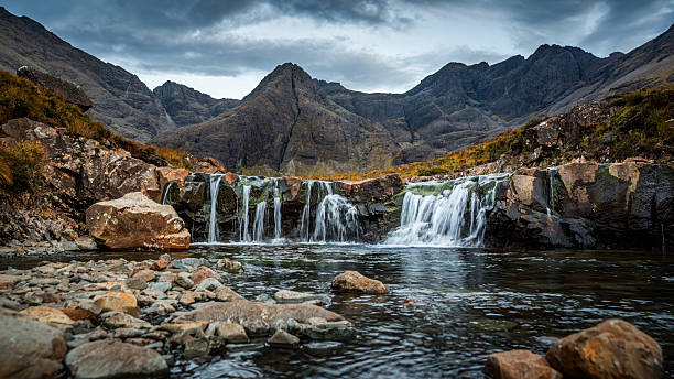 Fairy Pools on the Isle of Skye, a captivating series of waterfalls and crystal-clear pools set against the dramatic backdrop of the Cuillin Mountains. The foreground features a shallow, rocky stream leading to cascading waterfalls that spill over moss-covered rocks into pristine, calm pools. In the background, the rugged and towering Cuillin peaks dominate the scene, their jagged silhouettes adding an imposing and mystical quality. Scottish Highlands on the Isle of Sky in Scotland, United Kingdom.