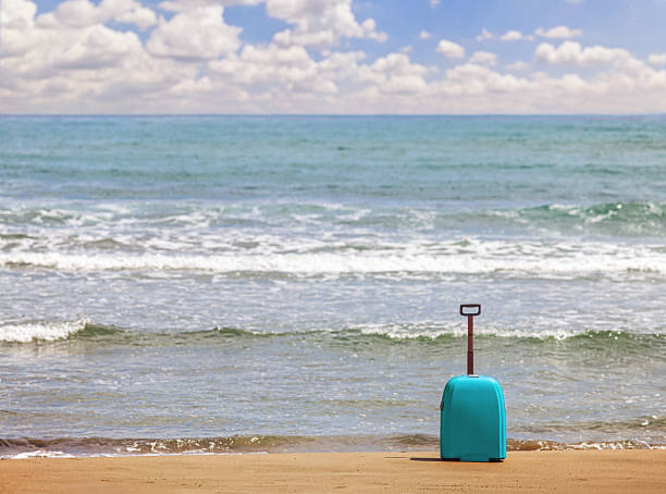 A bright teal rolling suitcase stands upright on golden sand against a backdrop of gentle ocean waves and a cloudy blue sky. This sunny composition captures the essence of summer holidays and international travel, making it ideal for booking agencies and tourism advertisements.