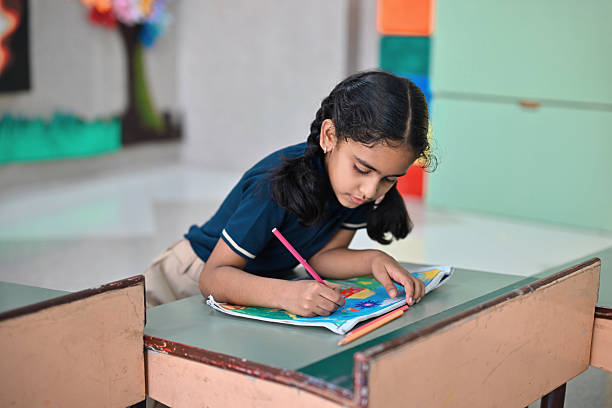 A young schoolgirl is seen intently coloring a worksheet while leaning over a desk in a vibrant classroom setting. She is focused and immersed in her artwork, using colored pencils. Childhood learning, creativity, and concentration in a school setting.