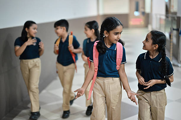 A group of cheerful elementary school students, wearing matching uniforms, walk together down a brightly lit school corridor. The children are engaged in lively conversation and laughter, some carrying backpacks, showing camaraderie and joy after class. The scene captures the essence of friendship, school life, and a positive learning environment in a modern educational setting.