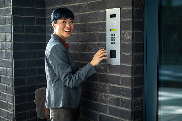 Asian businesswoman smiling while pressing doorbell on an intercom panel for secure access at a modern office building