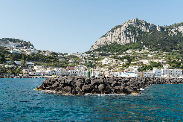 Capri, Italy - 04.29.2025: The end of the rocky pier in the Tyrrhenian Sea and the green light lighthouse structure. View from the sea and a view of the town in the background.