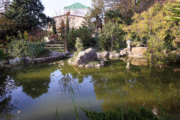 Tranquil Japanese-style garden featuring a calm pond surrounded by lush greenery, rocks, a wooden bridge, and traditional architecture in the background.