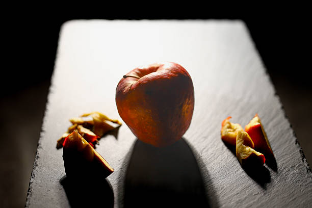 A whole apple is dramatically lit from behind, casting a shadow on a dark slate board. Surrounding the apple are pieces of cut peel, adding texture to the composition. The lighting emphasizes the apple's natural color and shape, creating a striking, minimalistic still life that highlights the raw beauty of natural produce.