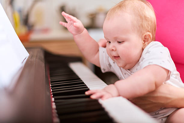 Baby girl, blonde, playing synthesiser with focused expression in bright room. Adult, supportive, hands visible. Warm lighting with blurred background
