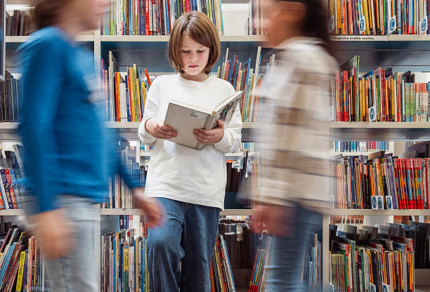 Young Caucasian boy standing in a library aisle, deeply engrossed in a book while people move around him, creating a dynamic scene of focused reading and bustling activity.