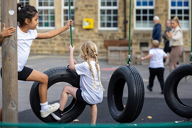A wide shot of two young school girls playing together on a climbing obstacle course. They are both wearing white school PE uniforms while climbing on black rubber tyres that are attached to the obstacle course.Videos are available similar to this scenario.