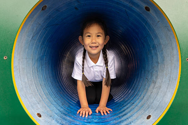 A medium shot of a young girl sitting inside of a play tunnel. She is wearing a white top and black pants as she is at school and taking part of a PE lesson. The young gilr is smiling while looking directly at the camera.Videos are available similar to this scenario.