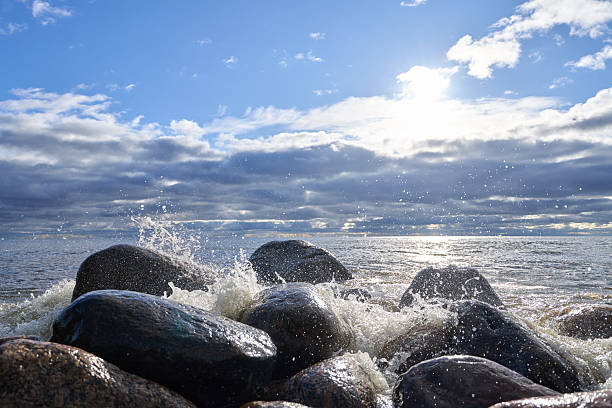 Detailed photograph capturing water splashes hitting rocky shore with sunlit reflections across sea surface, layered clouds and blue sky in background, taken in natural daylight.