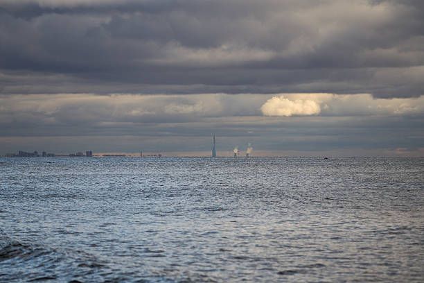 Wide seascape showing calm water surface leading to distant city skyline with tall modern tower, captured under dark cloudy sky and soft horizon light during daylight hours.