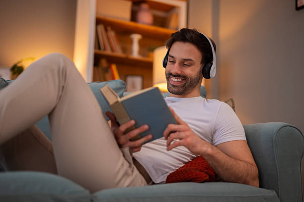 istock Smiling man wearing headphones reading book relaxing on couch 2248394740