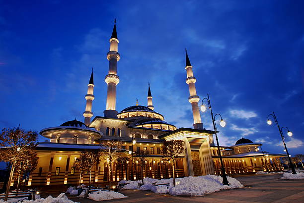 Ankara, Turkey - January 22, 2016 :A powerful, low-angle night photograph capturing the Millet Mosque (Millet Camii) in Ankara, Turkey, a prominent modern landmark built in the Ottoman-Turkish architectural style. The mosque is brilliantly illuminated in warm golden light against the deep blue hour sky. Patches of snow in the foreground and around the exterior lamps add a seasonal context, emphasizing themes of faith, grandeur, national identity, and winter serenity in the capital city.