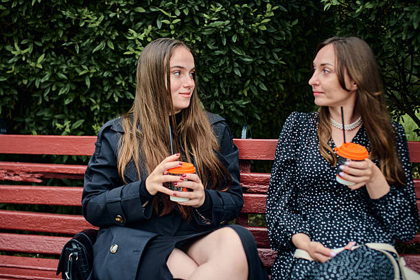 Young girlfriends sharing coffee and gossip on park bench with Greenery background