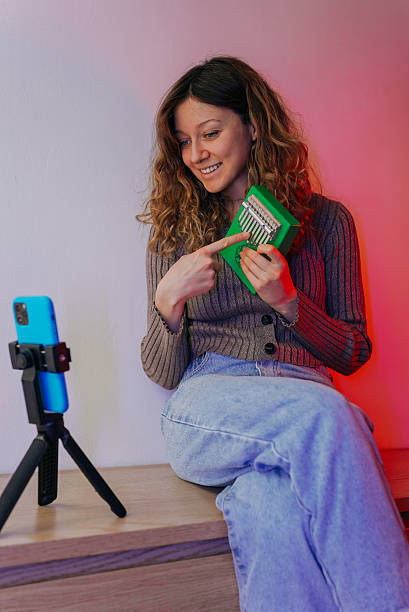 istock Female musician recording kalimba tutorial using smartphone mounted on tripod, seated on wooden surface, gesturing toward instrument, smiling while producing social media content 2248403437