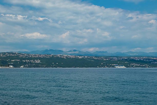 A panoramic view of the Croatian shore. There are some towns located on the shore of the Mediterranean Sea. Calm surface of the sea. Stony beach. Green hills in the back. Sunny day with puffy clouds
