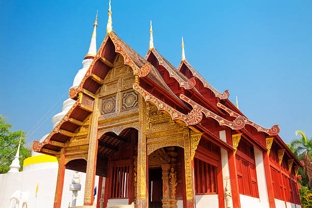 Thai temple building of Wat Phra Singh in Chiang Mai. Building is golden ornate decorated.  Behind is a white stupa.