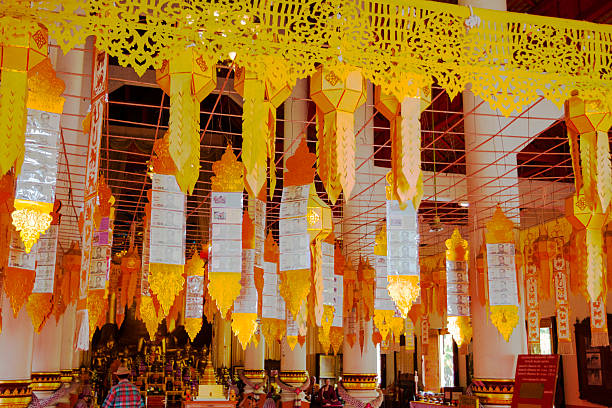 Hanging thai golden decor with bank notes in front of buddha statues inside of a temple of Wat Phra Singh in Chiang Mai city. A mature man is walking in backgrund below decor and close to many golden thai buddha statues. King Ananda Mahidol (Rama VIII), the older brother of King Bhumibol Adulyadej (Rama IX), gave the temple the status of Royal temple of the first grade.