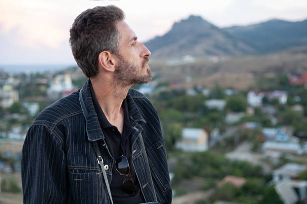A man with a short beard in a dark gray striped denim jacket in profile at sunset over mountain peaks and settlements