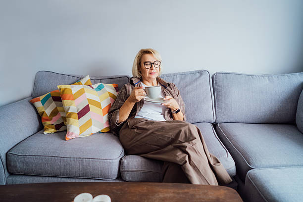 istock Middle aged smiling woman sitting on sofa and looking away while drinking coffee or tea. Mature senior woman relaxing after housekeeping, home cleaning. Portrait of relaxed female resting at home. 2248411187