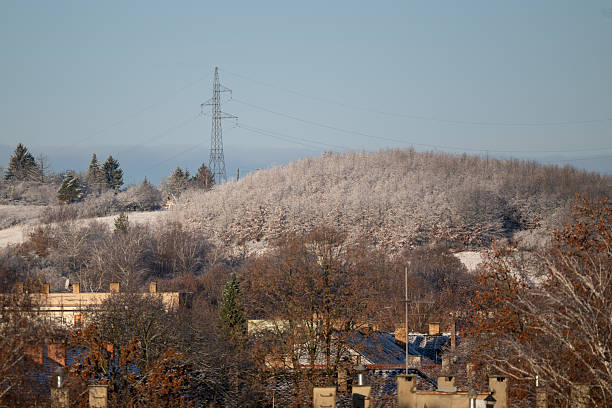A crisp winter scene showing a frost-covered hillside rising behind a small settlement with visible rooftops. Bare trees frame the view, while a tall power line tower stands against the clear blue sky, adding an industrial contrast. The soft morning light highlights the textures of snow and foliage.