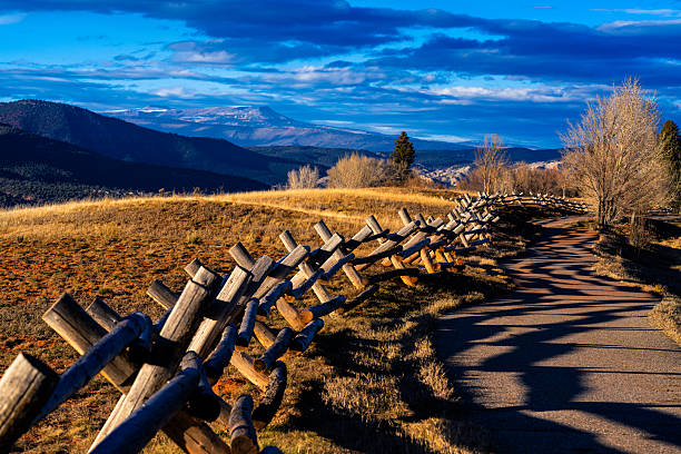 Scenic rural mountain area with wood fence running along recreation path casting long shadows on path with distant mountain views during golden hour.