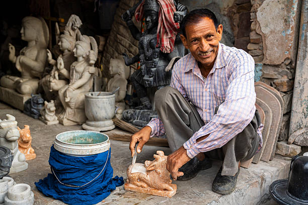A smiling artisan crouches in a small workshop, using a chisel to refine a small carved animal figure. He is surrounded by finished and in-progress stone and marble statues, sculpting tools, and buckets, conveying the atmosphere of traditional handcraft and skilled workmanship.