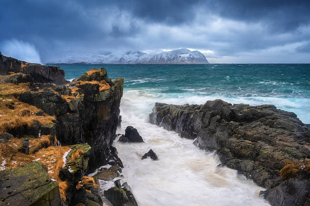 Wave impact on a rocky coastline with turquoise sea and snowy mountains in Lofoten Islands, Norway. Dynamic coastal landscape with cliffs, winter colors and rough water. Dramatic scenery with rocks