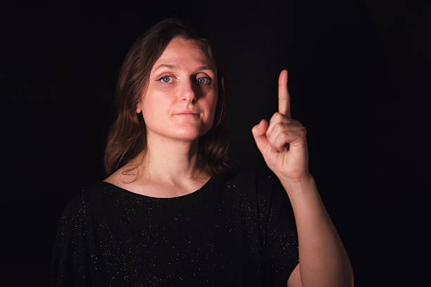 Caucasian woman pointing finger up, making a number one gesture. Serious expression with a dark background for attention or idea concept.