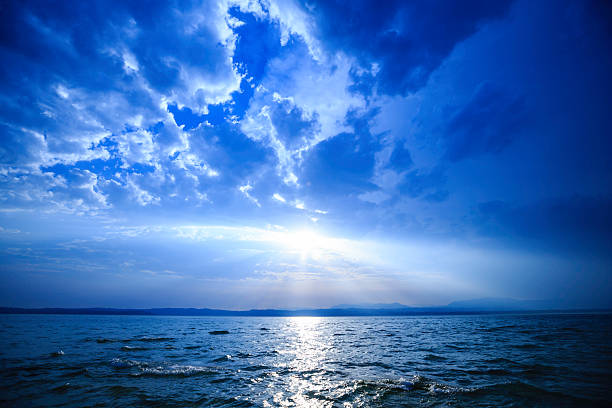 A tranquil seascape featuring a vast blue ocean, radiant sun piercing dense clouds, and distant land on the horizon. Perfect for travel, nature, and contemplative mood stock imagery.