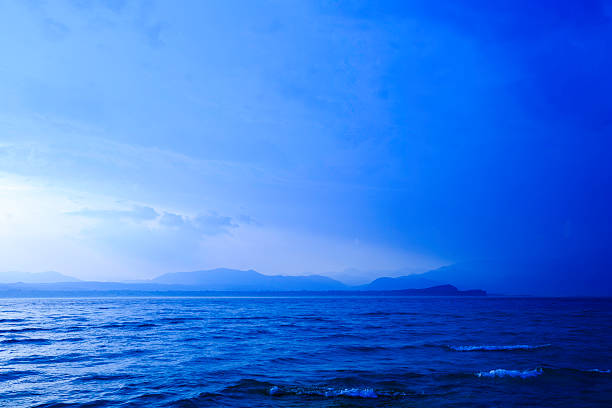 A serene seascape with calm blue waters extending to a hazy horizon of distant mountains beneath a wide, cloud-soft sky. Evokes peace, vastness, and nature’s beauty.