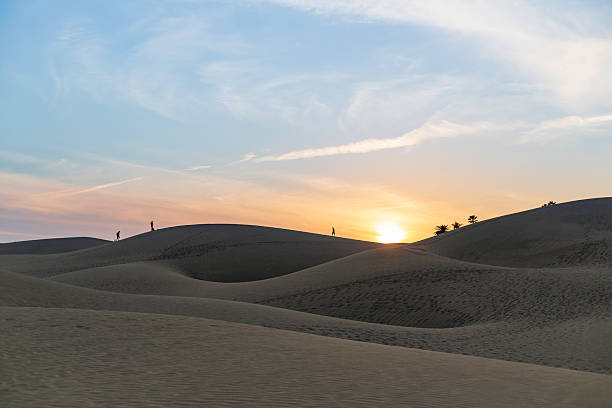 People walk along dune crests as the sun sets, creating silhouettes against a warm, orange horizon. The tranquil desert landscape conveys solitude, exploration, and the beauty of vast, open spaces.