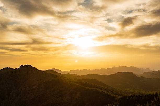 A warm, golden sunset washes over layered mountain ridges, creating a peaceful, contemplative mood. Silhouetted trees line the slopes as soft clouds drift across a bright sky.