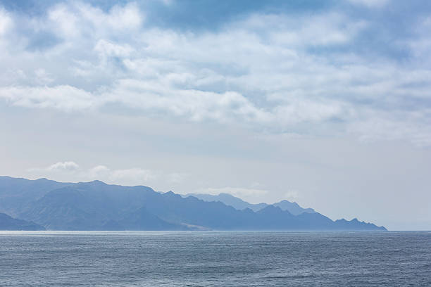 Calm ocean meets a rugged coastline with distant mountains under a soft, cloud-filled sky. A tranquil, expansive landscape that conveys peace and vast horizons.