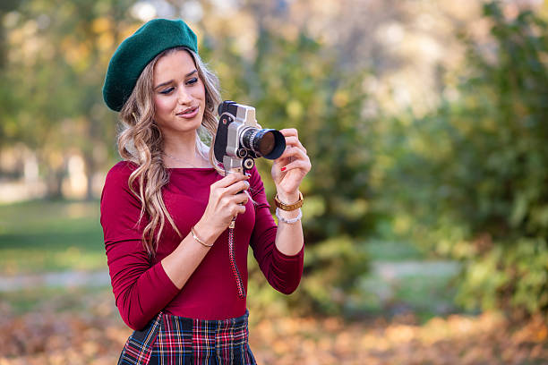 A beautiful young woman in a green beret filming video with retro video camera while standing in an autumn park.