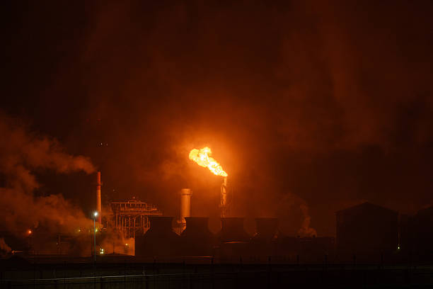 An industrial flare stack burns off excess gas with a large, bright orange flame and thick smoke against a dark night sky, highlighting energy production and potential environmental impact.
