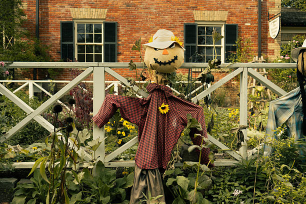 A person dressed as a scarecrow with a burlap mask, straw hat, and plaid shirt emerges from a stone well at night, raising arms with straw.