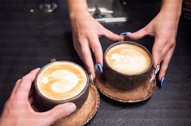 Two hands, one belonging to a barista serving, the other from a customer, reaching for two ceramic cups of cappuccino with beautiful latte art in a casual cafe setting