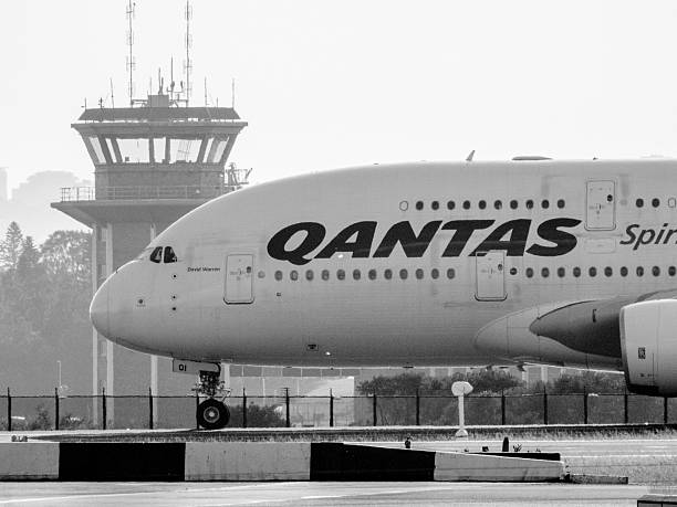 A Qantas Airbus A380-842 plane, registration VH-OQI, taxiing for departure from Sydney Kingsford Smith Airport as flight QF1 to London via Singapore. In the background is an air traffic control tower at the Airservices Australia complex on Botany Bay. This image faces west and the setting sun and was taken from Shep's Mound, Ross Smith Avenue, Mascot on a hot and sunny afternoon on 8 November 2025.