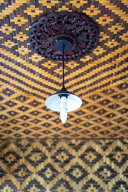 A low-angle shot of a ceiling featuring a strong, woven bamboo or rattan pattern in brown and light brown tones. A simple hanging pendant lamp with a modern bulb is attached to the center through a dark, decorative wooden carving (plafon/rosette). The texture is unique and traditional.