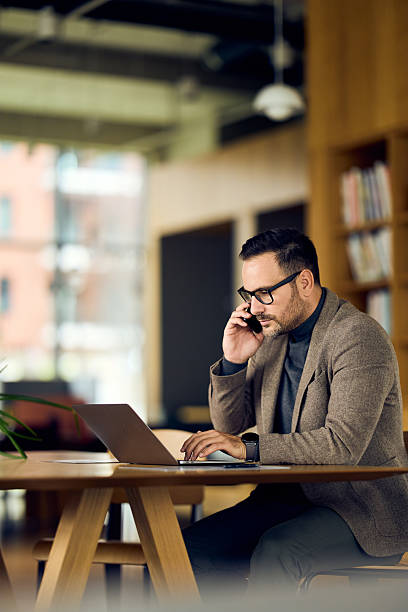 istock Focused Professional Man Working on Laptop While Talking on Phone in Modern Office 2248451979