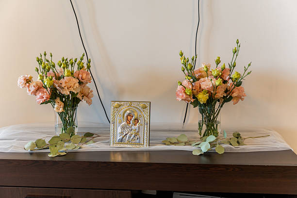 Vases of soft pink and yellow flowers are arranged on a wooden table beside a religious icon. The warm indoor setting highlights tranquility and beauty, perfect for reflection.