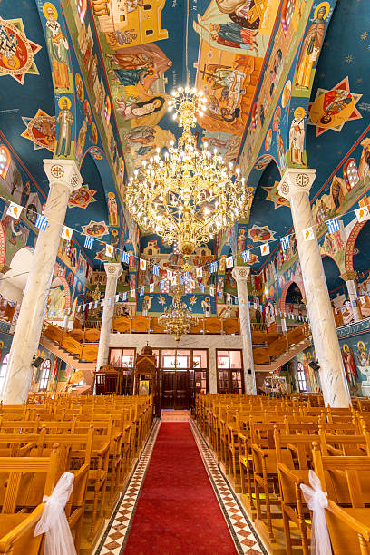Inside a beautifully decorated church, colorful murals adorn the ceiling and walls, while a large chandelier hangs over the red carpet. Rows of wooden chairs fill the space.