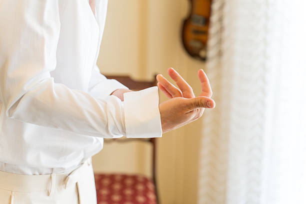 A groom is skillfully adjusting the cuffs of his white shirt, preparing for his wedding ceremony. The bright room exudes elegance with soft lighting and stylish decor.