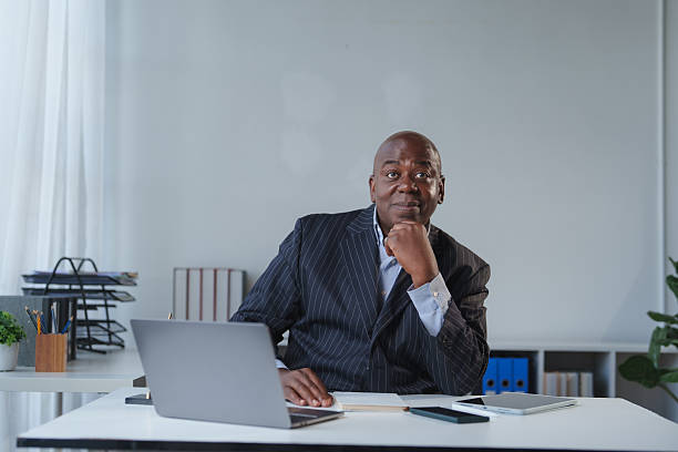Mature black businessman sitting at his office desk, looking at the camera and thinking, with a laptop and digital devices